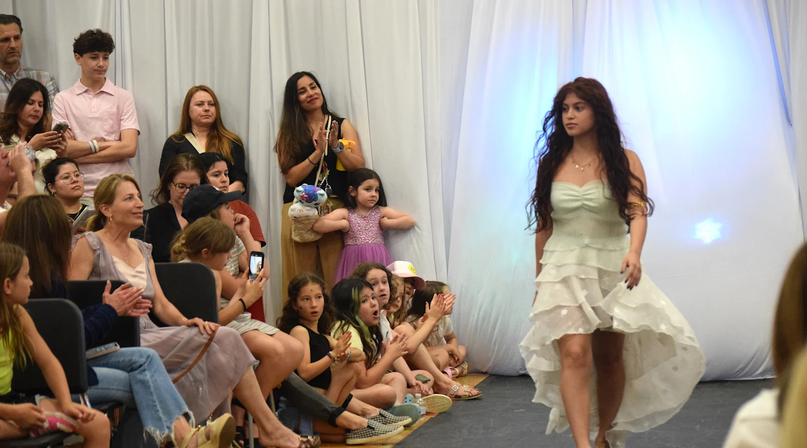 young students look on in awe as a high schooler walks down the runway in a white dress